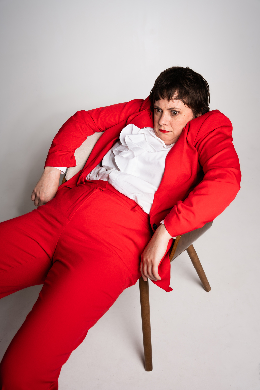 Emily Jeffers (white woman with short brown hair) sits slumped in a chair with an intense stare, wearing a bright red suit with a ruffled white dress shirt.